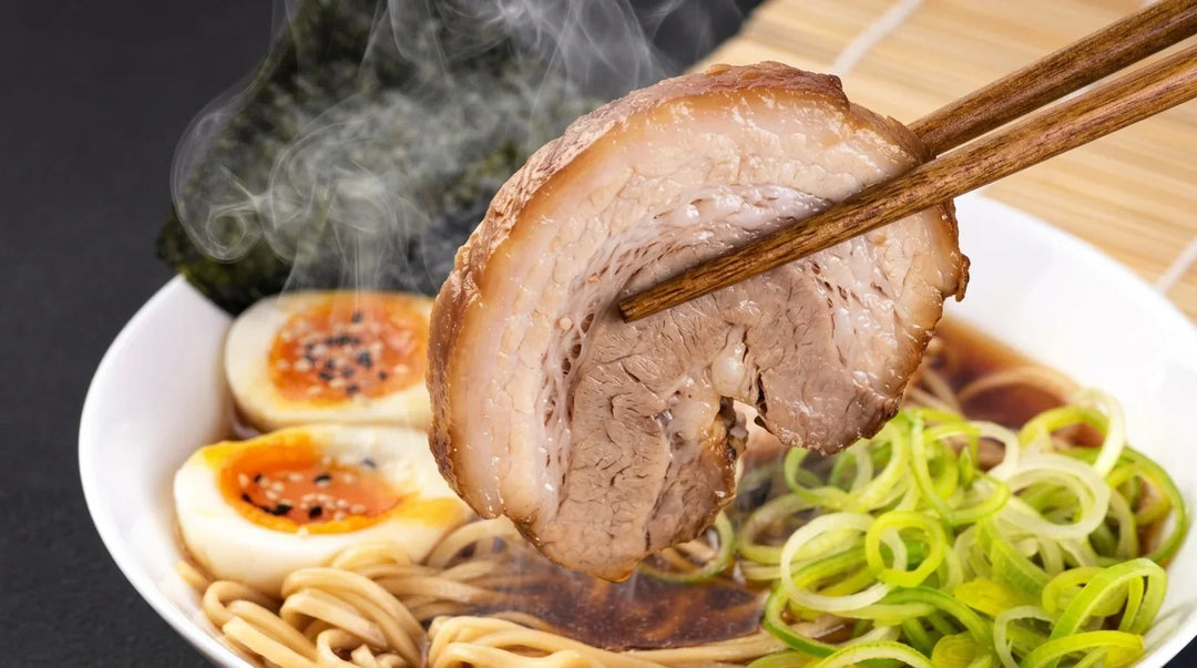 Ein Stück dampfendes Chashu-Schweinefleisch wird mit Holzstäbchen aus einer Ramen-Schale gehoben, die Nudeln, Brühe, zwei halbierte weichgekochte Eier mit orangefarbenen Dottern und schwarzen Sesamsamen, grüne Lauchzwiebelringe und ein Noriblatt enthält.