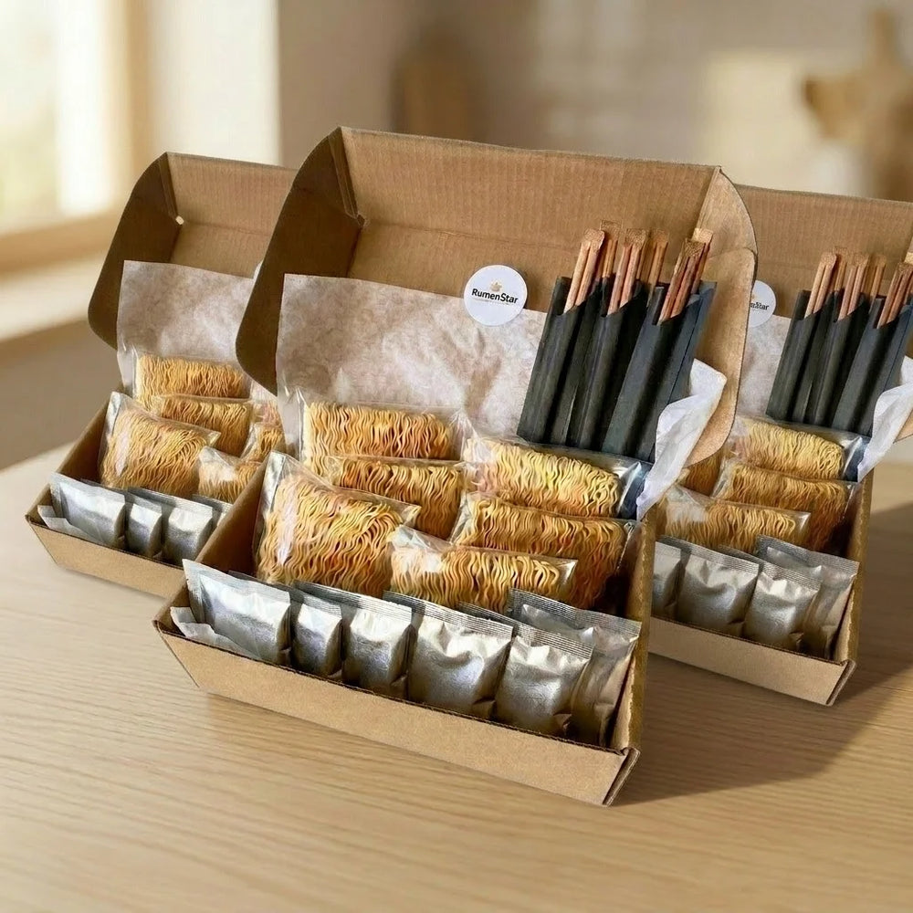 Drei offene braune Kartons stehen nebeneinander und zeigen den Inhalt von Instant-Ramen-Kits. Jeder Karton enthält mehrere einzeln verpackte, trockene Ramen-Nudeln und kleine silberne Gewürzsachets, die in zwei Reihen angeordnet sind. In jedem Karton befinden sich auch mehrere Paare Einweg-Essstäbchen aus Holz in schwarzen Papierhüllen. Ein Karton hat einen runden weißen Aufkleber mit dem Text "RumenStar" und einem Stern-Logo.