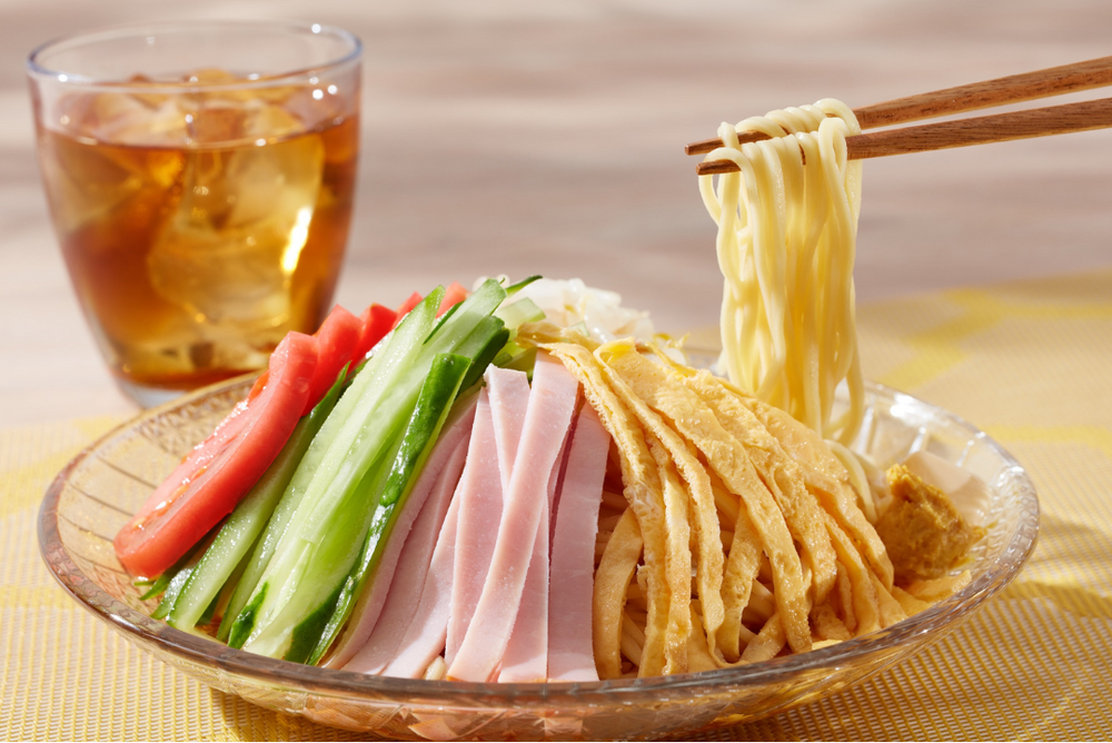 Kaltes erfrischendes Hiyashi Ramen Gericht mit RamenStar  Ramen Nudeln, mit Tomate, Schinken, Gurke und Omelette garniert. Staebchen ziehen Nudeln hoch. Im Hintergrund steht ein Glas kuehler Eistee mit Eis.