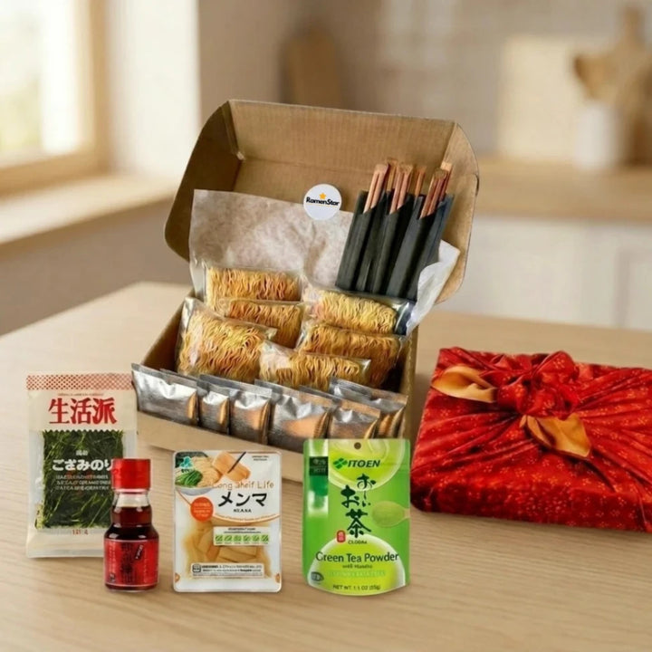Eine liebevoll zusammengestellte RamenStar Geschenkbox gefüllt mit frischen japanischen Ramen-Nudeln, Würzbeuteln und Essstäbchen. Daneben liegen Nori Algen, Menma-Bambussprossen, Chilisauce, Grüntee-Pulver und ein festlich in rotes Furoshiki verpacktes Geschenk, ideal für Liebhaber der japanischen Küche.