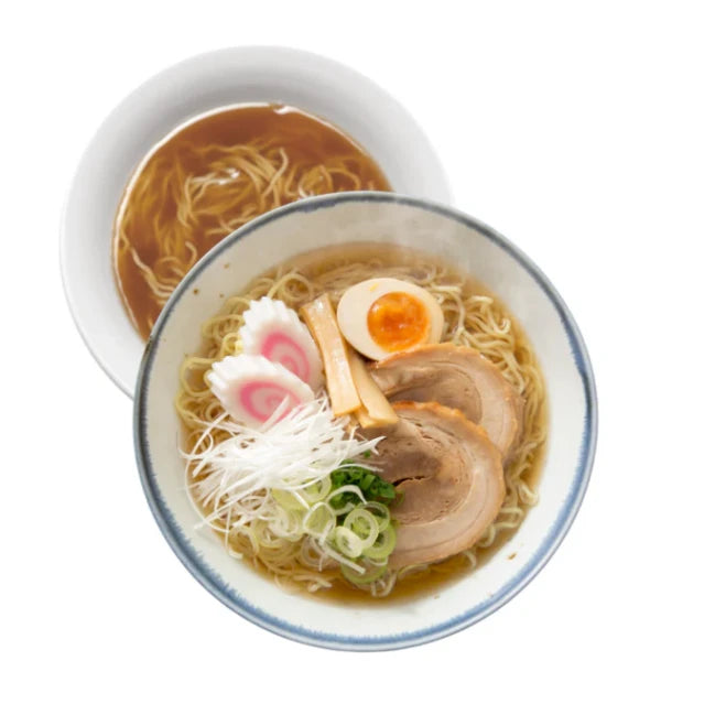 Eine dampfende Schale authentischer japanischer SHOYU-Ramen mit zarten Chashu-Schweinefleischscheiben, einem perfekt gekochten Ajitama-Ei, Narutomaki-Fischkuchen, Menma und frischen Frühlingszwiebeln. Eine weitere Nudelsuppe ist im Hintergrund.