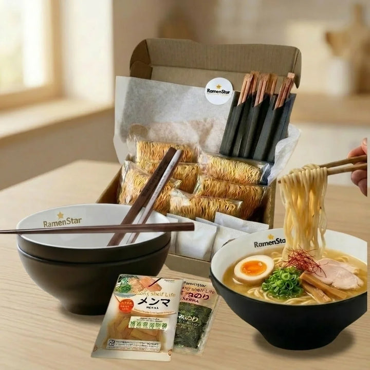 Eine geöffnete braune Box mit RamenStar-Logo, gefüllt mit mehreren Päckchen trockener Ramen-Nudeln, Gewürzpäckchen und verpackten Essstäbchen. Davor auf einem Holztisch stehen zwei schwarz-weiße RamenStar-Schalen. Die linke Schale ist leer mit Essstäbchen, die rechte Schale enthält fertige Ramen mit goldener Brühe, Nudeln, einem halbierten Ei, Fleischscheiben, Lauchzwiebeln, Bambussprossen und Chilifäden. Eine Hand hebt Nudeln mit Essstäbchen aus der gefüllten Schale. Zwei flache Päckchen mit Menma &amp; Nori.