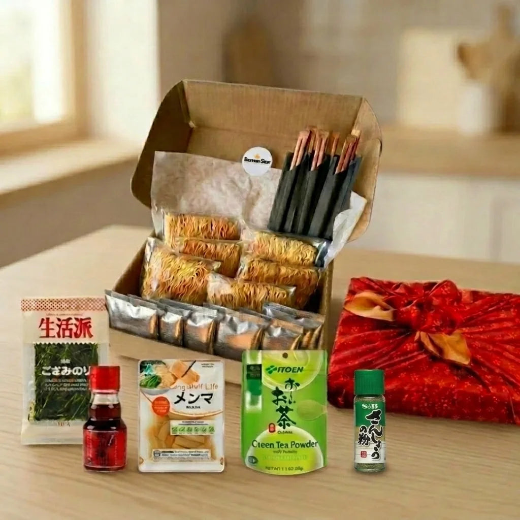 Eine liebevoll zusammengestellte RamenStar Geschenkbox gefüllt mit frischen japanischen Ramen-Nudeln, Würzbeuteln und Essstäbchen. Daneben liegen Nori Algen, Menma-Bambussprossen, Chilisauce, Grüntee-Pulver, Sansho Pfeffer und ein festlich in rotes Furoshiki verpacktes Geschenk, ideal für Liebhaber der japanischen Küche.