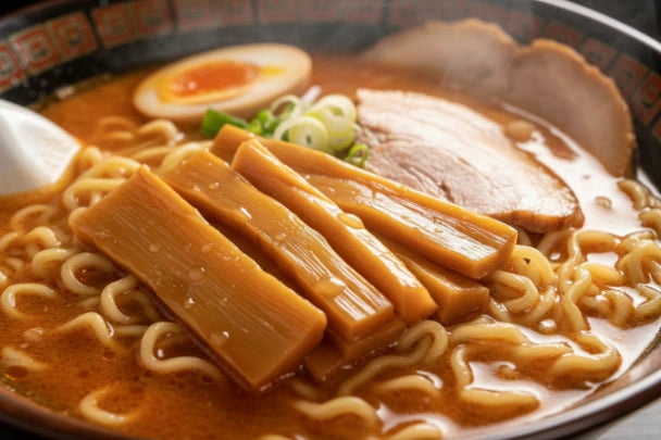 Nahaufnahme einer weißen Verpackung „Long Shelf Life“ Menma, japanische gewürzte Bambussprossen, ideal für Ramen. Das in Japan hergestellte Produkt liegt auf einem Holztisch, mit einer Schale ähnlicher Sprossen im dampfenden Hintergrund.