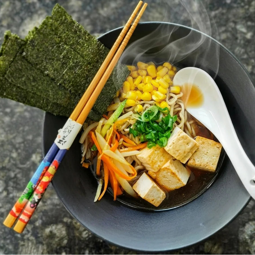Schüssel dampfender Ramen mit Nudeln, goldgelben Maiskörnern, gewürfeltem Tofu, juliennierten Karotten und Gurken, Sprossen und frischen Frühlingszwiebeln. Links daneben liegen zwei Blätter Nori und ein Paar bunte Essstäbchen. Eine weiße Keramikschale ist im Ramen platziert und leichter Dampf steigt aus der warmen Suppe auf.