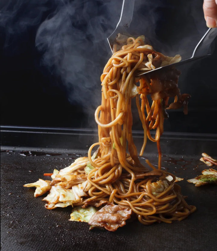Frisch zubereitete japanische Yakisoba-Nudeln mit zartem Schweinefleisch und knackigem Weißkohl werden dampfend von einer heißen Kochplatte gehoben. Das appetitliche Gericht glänzt und ist bereit zum Genuss.