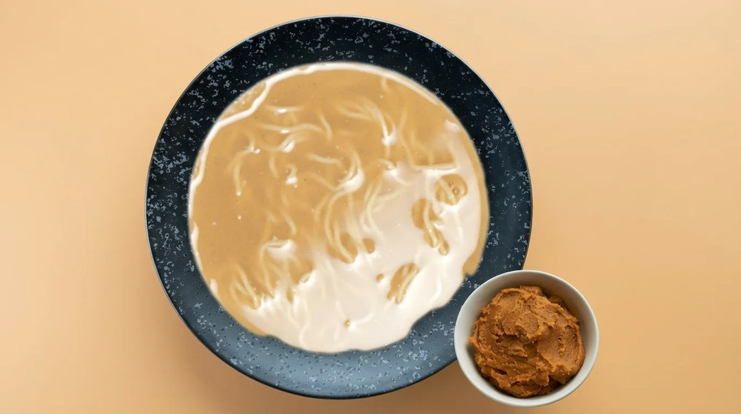 Dunkelblaues Geschirr mit Nudeln in hellbrauner Miso Suppe, daneben eine kleine weisse Schale mit brauner Miso Paste, auf beigem Hintergrund.