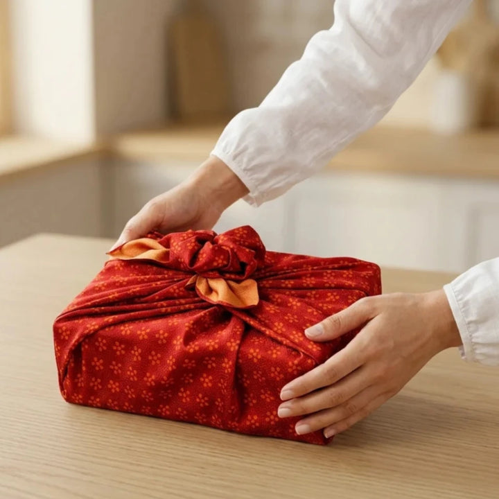 Hände in weißem Hemd platzieren ein festlich in rotem Furoshiki-Stoff mit goldenen Mustern und dekorativem Knoten verpacktes Geschenk auf einem Holztisch. Die nachhaltige, traditionelle Verpackung wirkt achtsam und persönlich.