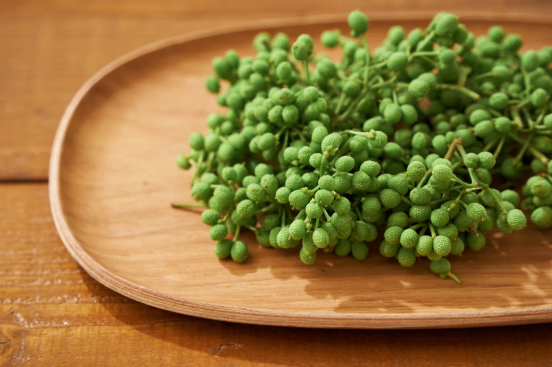Frische grüne Szechuan-Pfefferbeeren liegen dekorativ auf einem hellen, ovalen Holzteller. Die kleinen, knorrigen Beeren mit ihren Stielen sind vor einem warmen Holzhintergrund arrangiert und betonen die Lebendigkeit dieses einzigartigen asiatischen Gewürzes.