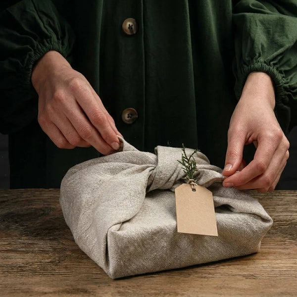 Eine Person mit dunkelgrüner Kleidung bindet ein Geschenk von Ramenstar, das in ein beigefarbenes aus Leinenstoff bestehendes japanisches Furoshiki Geschenktuch eingewickelt ist, auf einem Holztisch. Eine kleine grüne Zweigpflanze und ein leeres braunes Etikett sind dekorativ und edel am Stoffknoten befestigt.