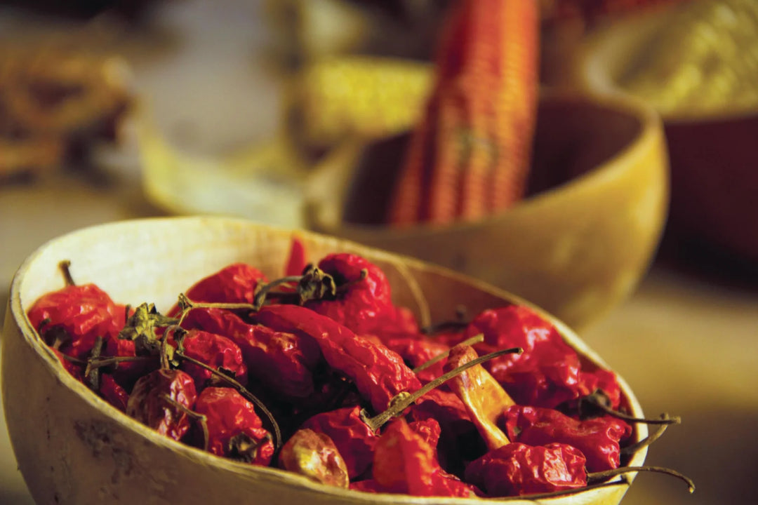 Rustikale Schale gefüllt mit leuchtend roten, getrockneten Chilischoten und Stielen. Im unscharfen Hintergrund sind weitere Ernteprodukte wie ein Maiskolben in einer ähnlichen Schale sichtbar, was eine warme, authentische Szene darstellt.
