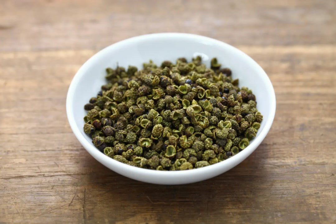 Eine weiße Schale gefüllt mit getrockneten, grünen Sichuan-Pfefferkörnern auf einem rustikalen Holztisch. Die einzigartigen Gewürzbeeren, viele davon leicht geöffnet, zeigen ihre charakteristische, raue Textur, perfekt für asiatische Gerichte.