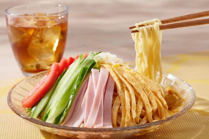 Appetitliches Hiyashi Chūka, japanischer kalter Nudelsalat mit frischen Gurken, Tomaten, Schinken und Omelettstreifen. Stäbchen heben die Ramen-Nudeln, begleitet von einem kühlen Eistee – ein perfektes Sommergericht.