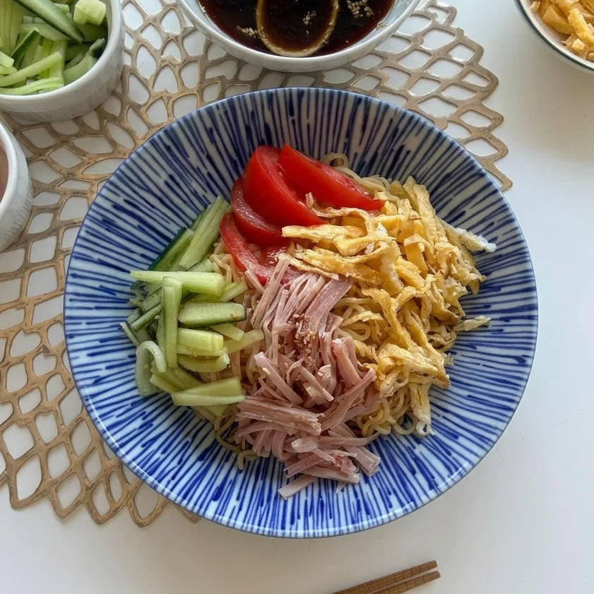 Hiyashi Chuka in einer blau-weiss gestreiften Schale, mit kalten Nudeln, drei Tomatenspalten, grünen Gurkenstreifen, hellrosa Schinkenstreifen und gelben Omelettenstreifen belegt. Daneben stehen eine Schale mit dunklem Soja-Dressing und Sesam, eine mit geschnittenen Gurken und eine mit geschnittenen Tomaten. Im Hintergrund ist eine weitere Schale mit Rühreistreifen sichtbar. Braune Essstäbchen liegen am unteren Bildrand auf einem hellbraunen Platzset.
