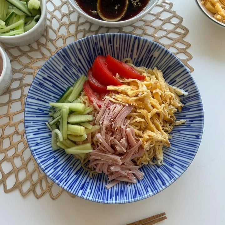 Hiyashi Chuka in einer blau-weiss gestreiften Schale, mit kalten Nudeln, drei Tomatenspalten, grünen Gurkenstreifen, hellrosa Schinkenstreifen und gelben Omelettenstreifen belegt. Daneben stehen eine Schale mit dunklem Soja-Dressing und Sesam, eine mit geschnittenen Gurken und eine mit geschnittenen Tomaten. Im Hintergrund ist eine weitere Schale mit Rühreistreifen sichtbar. Braune Essstäbchen liegen am unteren Bildrand auf einem hellbraunen Platzset.