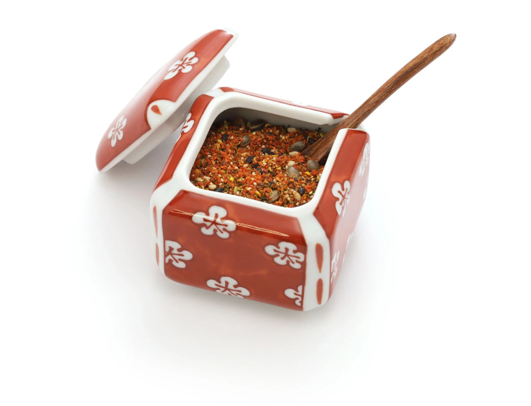 Ein eleganter, roter Keramik-Gewürzbehälter mit weißem Blütenmuster, gefüllt mit farbenfroher japanischer Shichimi Togarashi Gewürzmischung. Ein kleiner Holzlöffel ruht in den Gewürzen, ideal für die Verfeinerung asiatischer Gerichte.