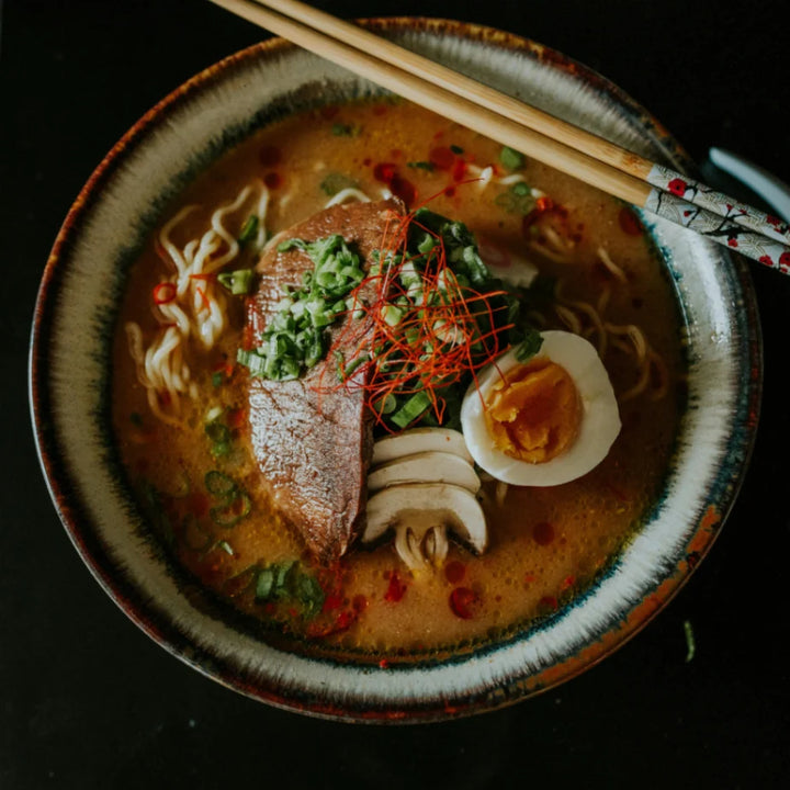 Eine köstliche japanische Ramen-Nudelsuppe mit herzhafter Brühe, zartem Rindfleisch, einem marinierten Ei, frischen Pilzen und Lauchzwiebeln, garniert mit roten Chilifäden und Essstäbchen auf dunklem Hintergrund.