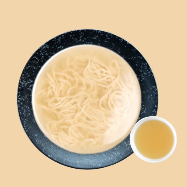 Draufsicht auf eine minimalistische Nudelsuppe mit heller Brühe und dünnen Nudeln in einer dunkelblauen, gesprenkelten Keramikschale, daneben eine kleine Tasse mit zusätzlicher klarer Shio Brühe auf beigem Hintergrund.