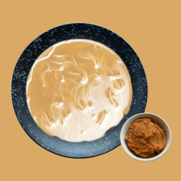 Dunkle Schüssel mit cremigem, hellbraunem Miso Nudelsuppe. Daneben eine kleine weisse Schale mit dicker, orangebrauner Miso-Paste asl Geschmacksbeispiel.