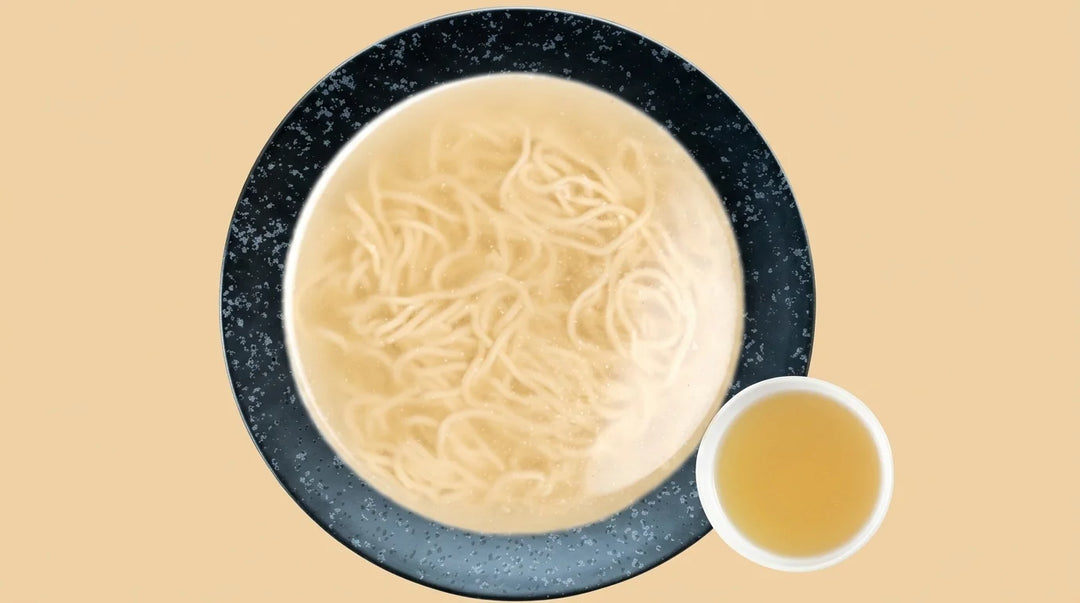 Dunkelblaue Schale mit weissen Nudeln in klarer, heller Brühe und eine kleinere, weisse Schale mit goldgelber Flüssigkeit daneben auf einem beigen Hintergrund, von oben gesehen.