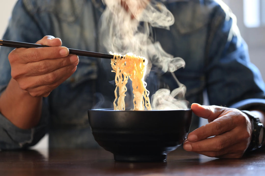 Nahaufnahme einer Person, die mit Essstäbchen dampfende goldene Nudeln aus einer schwarzen Schüssel hebt, mit viel weißem Dampf auf einem dunklen Holztisch. Eine herzhafte asiatische Mahlzeit.