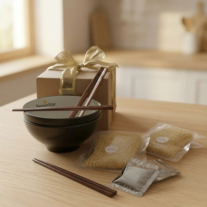 Ein brauner Geschenkkarton mit goldener Schleife steht hinter einer schwarzen Ramenschale mit dem "RamenStar"-Logo und braunen Essstäbchen. Auf dem hellen Holztisch daneben liegen zwei Packungen getrockneter Ramen-Nudeln und zwei kleine silberne Gewürztütchen.