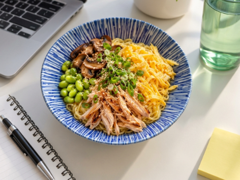 Kalte Ramen mit gebratenen Pilzen, Edamame, Poulet und Sesam am Schreibtisch neben einem Laptop, Meal Prep fürs Büro.
