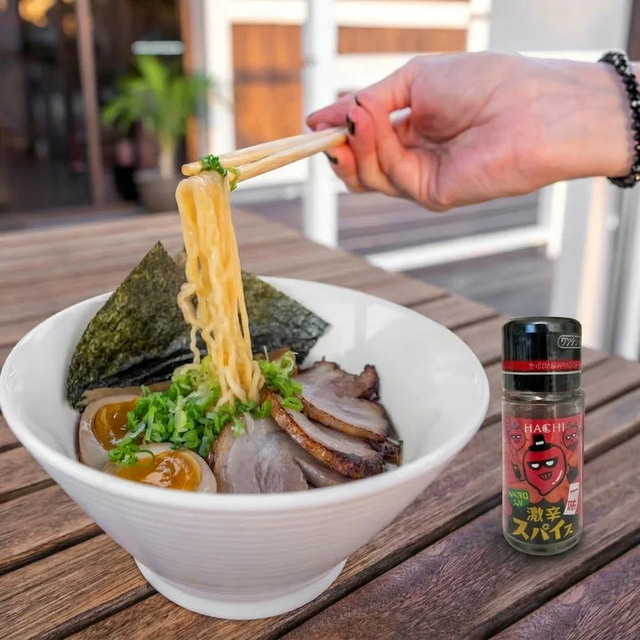 Eine Hand hebt mit Holzstäbchen goldgelbe Ramen-Nudeln aus einer weißen Schale. Die Schale enthält Ramen mit geschnittenem Schweinefleisch (Chashu), zwei halbierten, marinierten Eiern, gehackten grünen Frühlingszwiebeln und zwei dreieckigen Nori-Blättern. Rechts neben der Schale steht eine kleine, rote Gewürzflasche der Marke Hachi Gekikara Spice. Alles steht auf einem hellbraunen Holztisch.