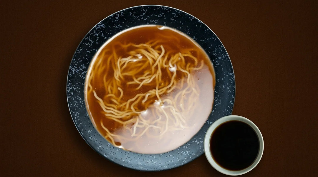 Ansicht von oben auf eine dunkelblaue, gesprenkelte Schale, gefüllt mit dünnen, hellbraunen Ramen-Nudeln in einer klaren braunen Shoyu Brühe. Rechts daneben steht eine kleine weisse Schale mit dunkler Sojasauce. Alles liegt auf einem braunen, strukturierten Stoffhintergrund.