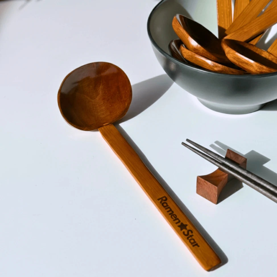 Glänzender, dunkelbrauner Holzlöffel mit 'Ramen Star' Gravur, daneben eine graue Schale mit weiteren Holzlöffeln und Essstäbchen auf einem Holzhalter. Perfekt für authentischen Ramen-Genuss.
