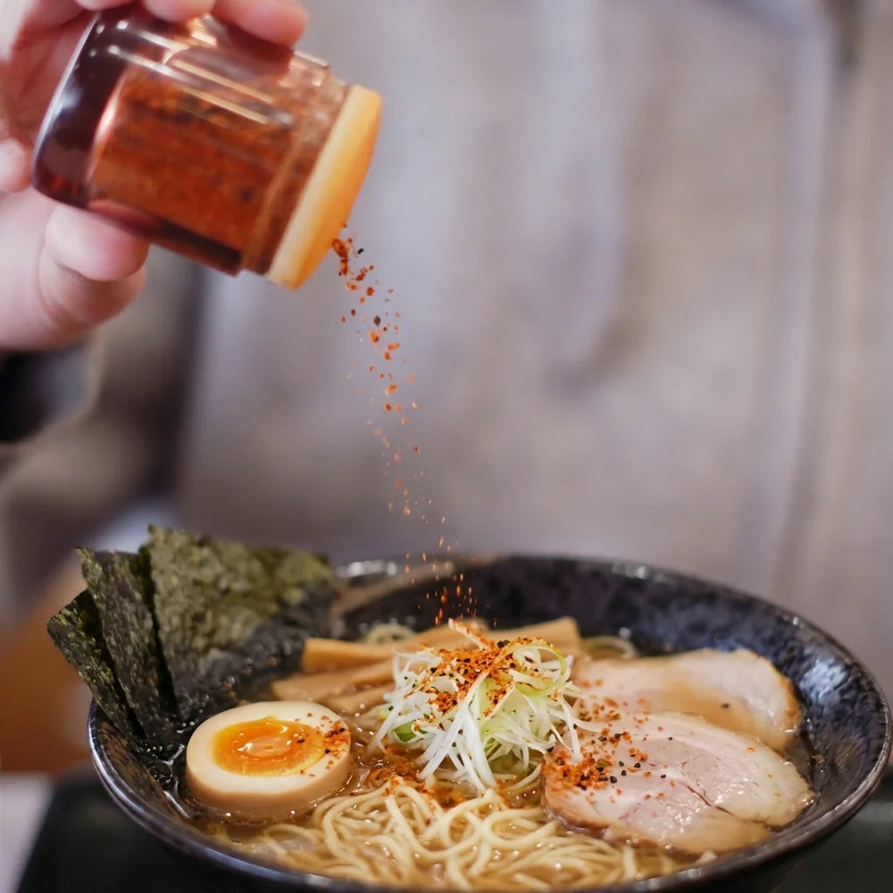 Eine Hand streut rote Chiliflocken aus einem Streuer über eine Schale mit Ramen. Die Ramenschale enthält Nudeln, Brühe, geschnittenen Chashu-Schweinebauch, ein halbiertes weichgekochtes Ei, Lauchzwiebeln, Bambussprossen und Nori-Algen. Einzelne Flocken sind in der Luft sichtbar, während sie auf das Gericht fallen.