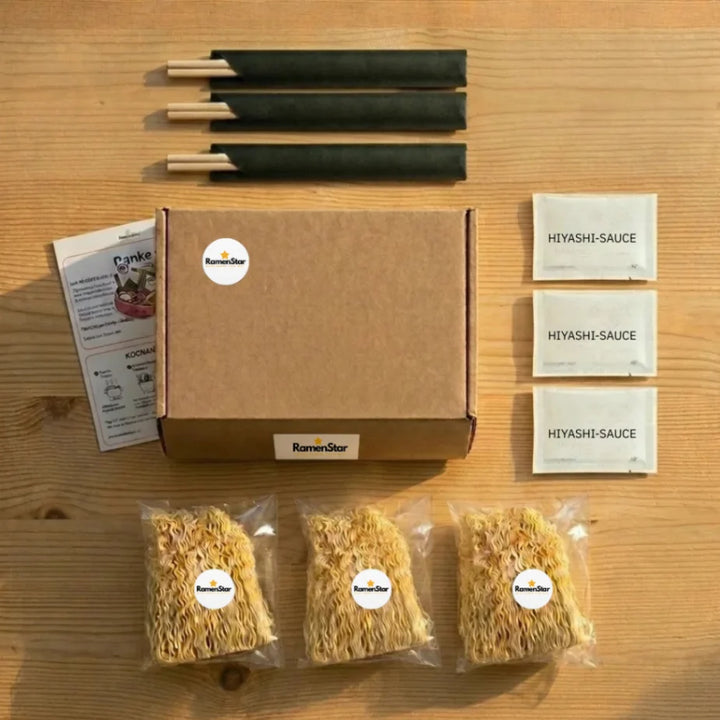 RamenStar Kochset-Inhalt, übersichtlich auf Holztisch präsentiert: Eine braune Box, drei Packungen Hiyashi-Sauce, drei Portionen trockener Ramen-Nudeln und drei Paar Essstäbchen. Perfekt für ein schnelles, authentisches japanisches Gericht.