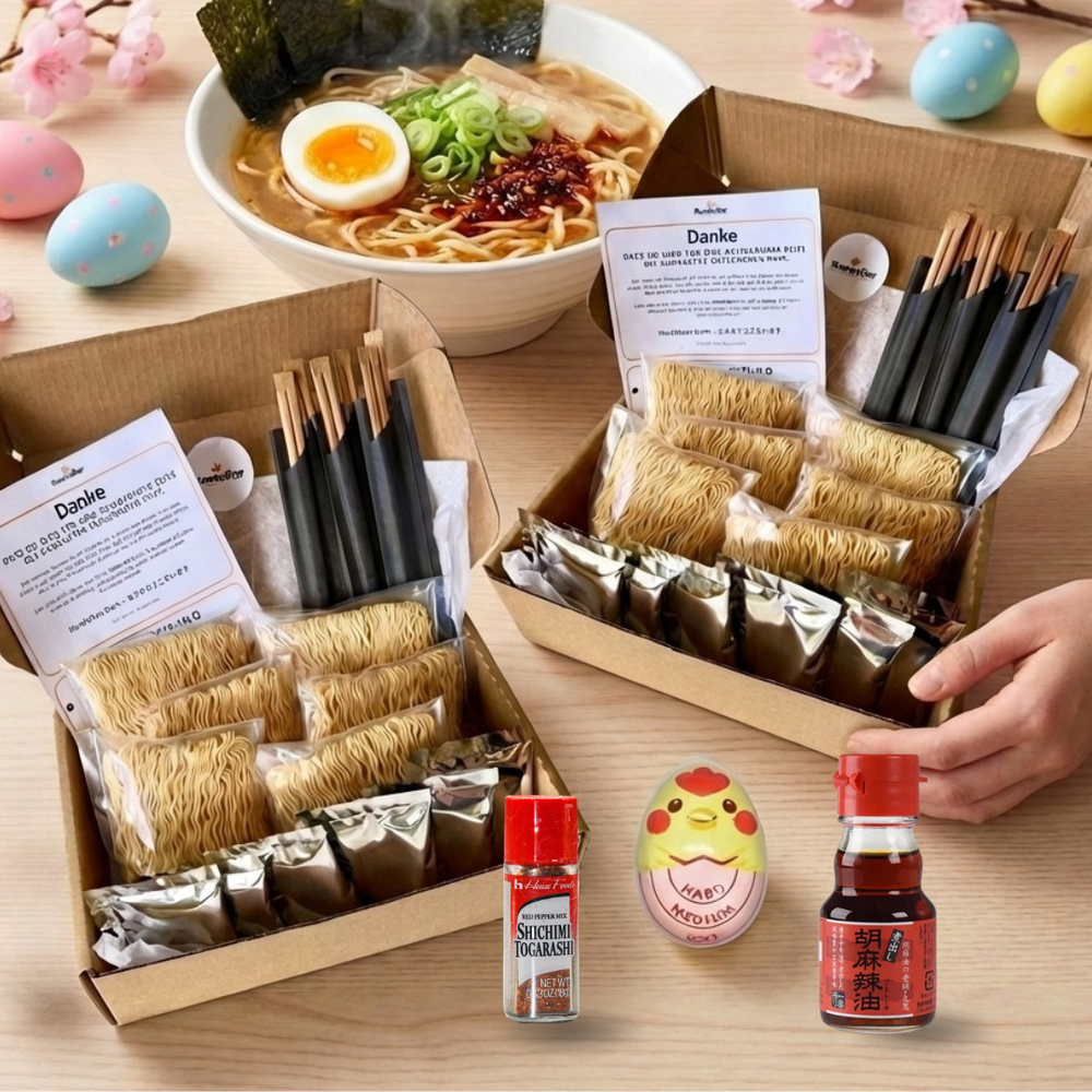 Zwei offene Kartons mit Ramen-Kochsets, gefüllt mit Nudeln, Soßenbeuteln und schwarzen Essstäbchen. Im Hintergrund steht eine dampfende Schüssel mit zubereitetem Ramen, garniert mit einem halbierten Ei, Nori, Frühlingszwiebeln und Chili. Auf dem Holztisch sind rosa Kirschblütenzweige, drei bunte Ostereier, eine Hühnchen-Eieruhr und zwei kleine Gewürzflaschen verteilt. Eine Hand hält einen der Ramen-Kartons.