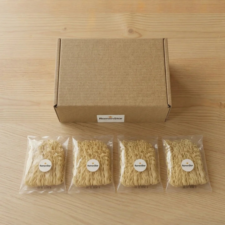 Brauner Versandkarton mit RamenStar-Logo und vier durchsichtigen Päckchen abgepackter Ramen-Nudeln auf einem hellen Holztisch.