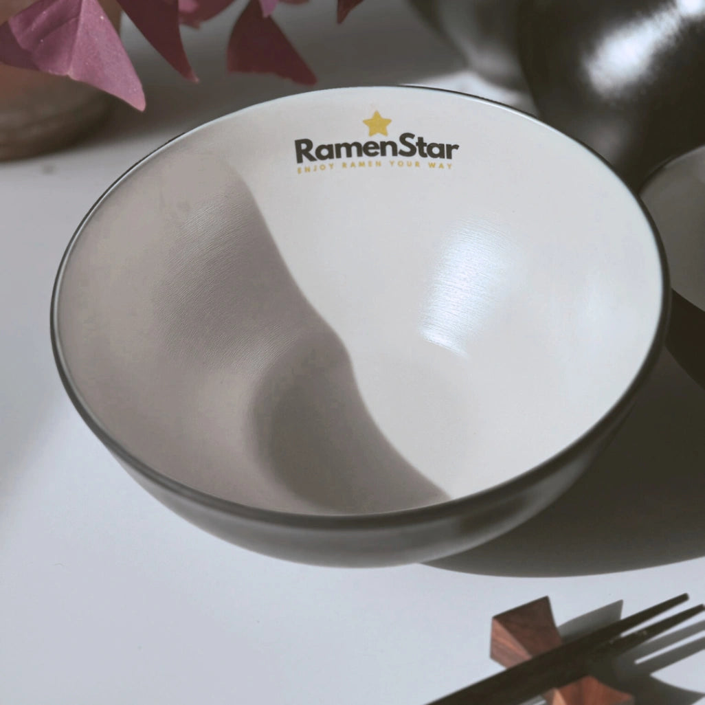 Eine hochwertige, minimalistische RamenStar Schale in Weiß und Dunkelgrau, mit dem goldenen RamenStar Logo und Slogan "ENJOY RAMEN YOUR WAY". Sie steht neben eleganten Essstäbchen, ideal für authentischen Ramen-Genuss zu Hause.