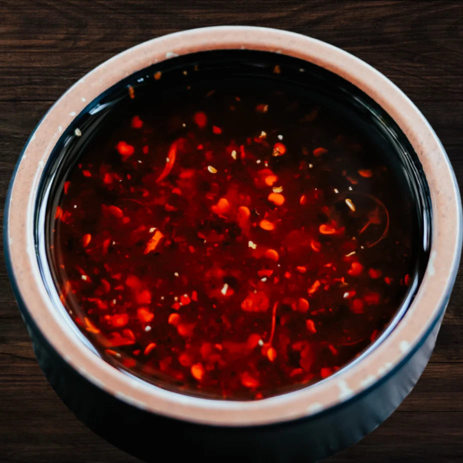 Eine Nahaufnahme einer dunkelroten, feurigen Chili-Öl-Sauce mit zahlreichen Chiliflocken und Samen, präsentiert in einer rustikalen Keramikschale auf einem dunklen Holztisch. Ideal als würzige Beilage für asiatische Gerichte.