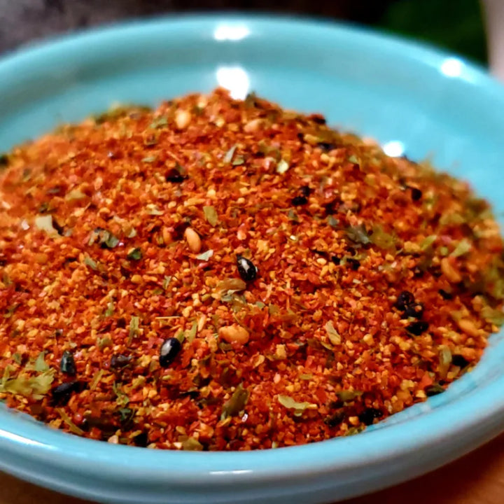 Nahaufnahme einer appetitlichen, leuchtend rot-orangefarbenen Shichimi Togarashi Gewürzmischung, angerichtet in einer hellblauen Schale. Die Mischung enthält Chiliflocken, grüne Kräuter, helle und schwarze Sesamsamen, ideal zum Verfeinern asiatischer Gerichte.