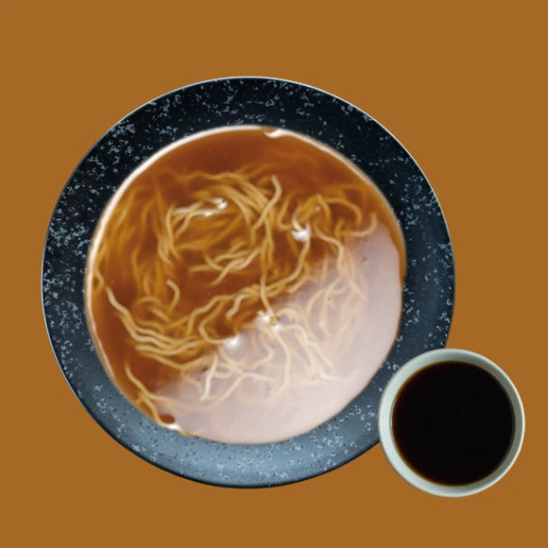 Appetitliche asiatische Nudelsuppe in dunkler Schale, von oben gesehen. Dünne Nudeln schwimmen in reichhaltiger brauner Shoyu Brühe, begleitet von einer kleinen Schale dunkler Sosse.