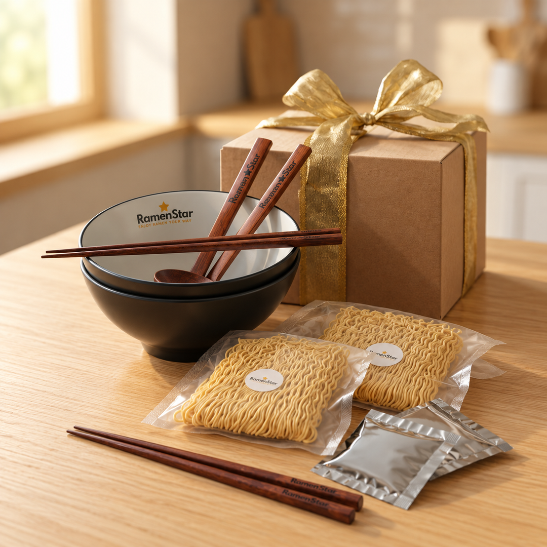 Zwei schwarze Ramen-Schalen, ineinander gestapelt, mit weissen Innenseiten und dem "RamenStar"-Logo. Darauf liegen braune Essstäbchen und ein Löffel. Vor den Schalen liegen zwei Packungen Instant-Ramen-Nudeln und zwei Gewürzpäckchen auf einem hellen Holztisch. Ein weiteres Paar brauner "RamenStar"-Essstäbchen liegt vor den Nudeln. Im Hintergrund steht ein braunes Geschenkpaket mit goldener Schleife.